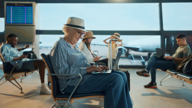 Senior woman in hat works on laptop while seated in a busy airport lounge, surrounded by diverse travelers — capturing modern mobility, remote work, and active lifestyle during travel.