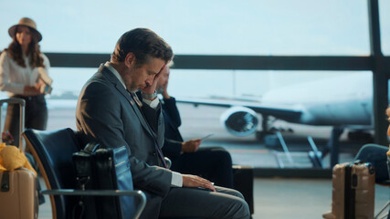 Business traveler sits with head down in airport lounge, appearing tired or overwhelmed while...