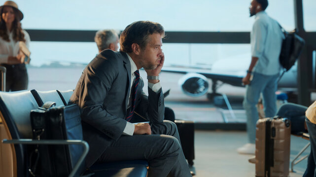 Worried businessman in a suit sits alone at the airport gate, lost in thought while waiting for his flight, reflecting the stress and uncertainty of corporate travel.