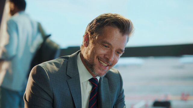 Close-up of smiling businessman in suit sitting at the airport, enjoying a lighthearted moment during travel — relaxed, professional atmosphere with soft daylight and terminal in the background.
