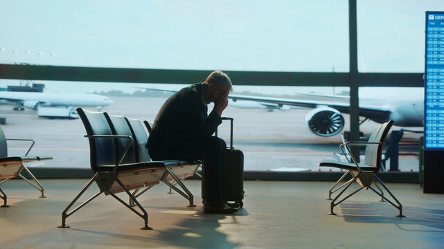 Stressed Business traveler sitting alone in airport lounge with head in hands, showing stress or frustration while waiting with luggage, airplane visible through large window on cloudy day