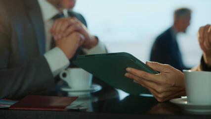 Close-up of hands holding digital tablet during business meeting at airport café, with coffee on the table and blurred professionals in background — symbolizing mobile work, collaboration, and travel 
