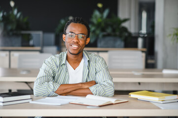 Smiling university student sitting at desk with crossed arms and books around