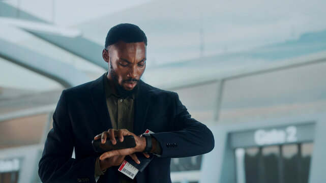Focused businessman checks the time on his watch while holding passport and boarding pass in airport terminal — illustrating time pressure, travel readiness, and efficiency during a business trip.