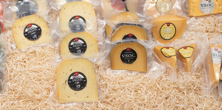 Various vacuum-sealed cheese wedges and wheels, primarily "Amanti" and "V.S.O.C." brands, arranged on bed of shredded straw material for display. Ukraine, Zhytomyr, July, 21, 2025