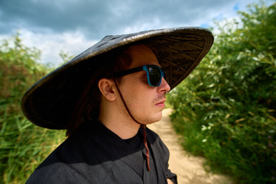 Man wearing a wide-brimmed hat walking along a nature path surrounded by greenery on a partly cloudy day