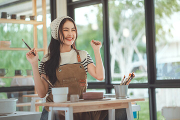 Joyful Asian female potter smiling brightly while holding a paintbrush, preparing to paint a...