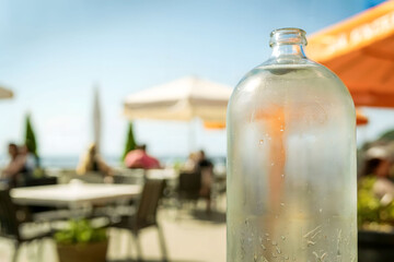 Frosted Glass Bottle of Chilled Water in Focus at Outdoor Café on Sunny Day with Blurred Background of People and Umbrellas
