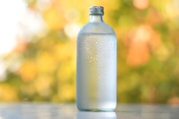 Frosted Glass Bottle with Condensation on Reflective Surface Against Warm, Blurred Vibrant Background