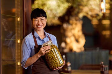 Smiling Female Shopkeeper Holding a Jar of Green Olives in Her Store