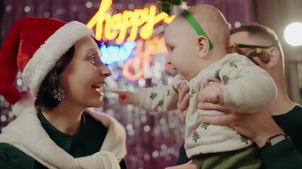 Joyful family celebrating new year with their baby in a festive atmosphere. The mother wears a Santa hat while holding the smiling baby. Illuminated 'Happy New Year' sign in the background.