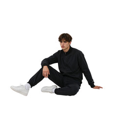 Young man in stylish black tracksuit and white sneakers sitting casually, looking directly at the camera with a relaxed posture, conveying modern fashion.