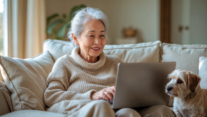 Smiling senior Asian woman works on her laptop with her dog at home on the sofa