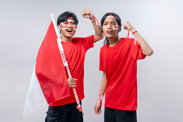 Two proud young men dressed in red T-shirts pose energetically with fists raised and Indonesian flag in hand, wearing face paint and headbands to celebrate Indonesia’s Independence Day.
