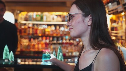 Beautiful woman guest zipping to taste in Christmas blue Lagoon cocktail with good mood sitting on luxurious counter bar green neon snow Christmas pine tree festive theme on wine background. Vinosity.
