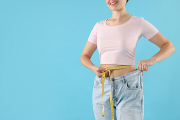 Woman with measuring tape wearing old jeans after weight loss on light blue background, closeup....
