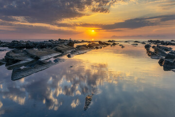 Sunset over the ocean with rocks in the foreground and calm waves, creating a scenic seascape.