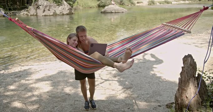 Couple enjoys online yoga class in nature by the river, using laptop in hammock. Exercising and stretching.