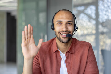 A smiling man wearing a headset waves during a video call, conveying positivity and engagement. He...
