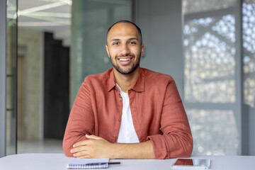 A smiling man sits at a table, appearing confident and relaxed. The setting suggests a modern office or workspace.