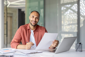 A smiling man sits at a desk, holding documents with a laptop nearby. He appears to be reviewing...