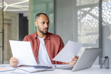 A focused businessman reviewing paperwork, studying documents in a modern office environment, next to a laptop and other work-related items.
