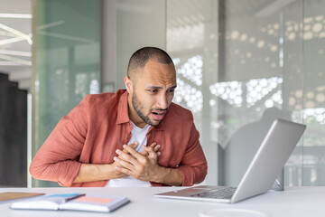A man in a modern office, shocked by what he sees on his laptop, clutches his chest in disbelief.