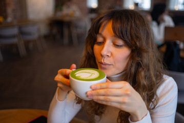 Young woman savoring the rich aroma and delightful taste of a warm matcha latte in a cozy cafe,...