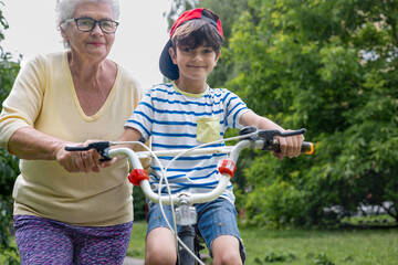 Obraz premium Grandmother Guides Grandson Learning to Ride Bicycle with Training Wheels. Family Bonding, Child Development, Active Summer Outdoor Activity. Copyspace