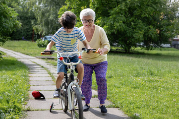 Grandmother Guides Grandson Learning to Ride Bicycle with Training Wheels. Family Bonding, Child...