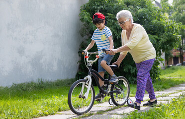 Grandmother Guides Grandson Learning to Ride Bicycle with Training Wheels. Family Bonding, Child...