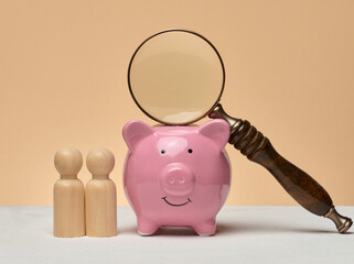 Piggy bank, wooden human figures, above the piggy bank there is a magnifying glass. The composition symbolizes financial analysis, investments or search for funds.