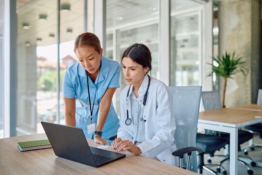 Female doctor and nurse analyzing medical data on a laptop, working together in a bright hospital office