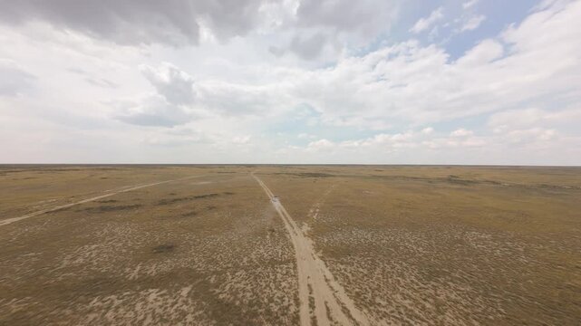 Aerial video of a white SUV driving fast through the dry yellow steppe of Ustyurt Plateau in Mangystau, Kazakhstan. Shot from drone in cinematic motion