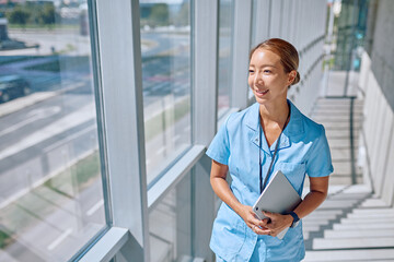 Young asian female doctor wearing blue uniform holding tablet and walking in modern hospital corridor next to large windows