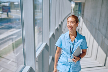 Smiling asian female doctor walking in modern hospital corridor holding tablet, healthcare and...