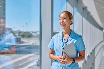 Young asian female doctor wearing blue uniform holding tablet and looking out the window in a modern hospital during a sunny day