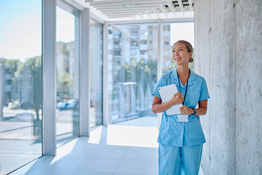 Portrait of a smiling asian female doctor in blue scrubs holding a tablet while walking in a modern hospital corridor