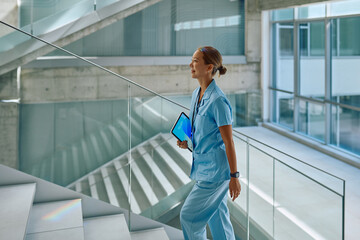 Smiling female doctor in blue scrubs holding digital tablet walking up stairs in modern hospital building