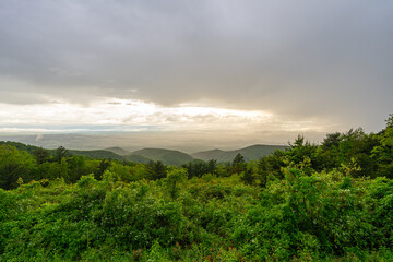 Obraz premium Panoramic view of rolling green mountains and valleys at sunset in Shenandoah National Park, Virginia, with dramatic storm clouds and soft golden light breaking through the sky.