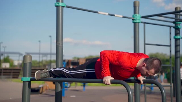 Man in red shirt doing dips on parallel bars at outdoor gym. He looks down.