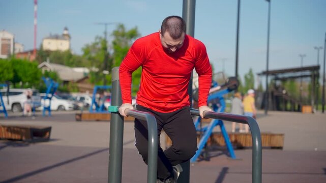 Man in red shirt doing dips on parallel bars at outdoor gym. He looks down.