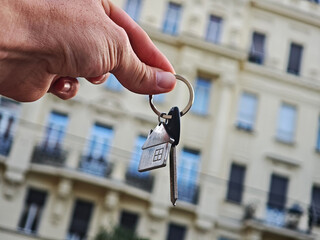 Hand holding house key with keychain in front of modern skyscraper, symbol of property investment