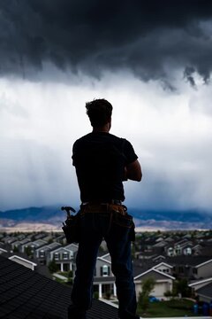 roofer with his arms crossed, silhouette, standing proudly atop the rooftop of a house, sky churns an ominous gray storm cloud on horizon, roofing company advertisement, roof repair social media
