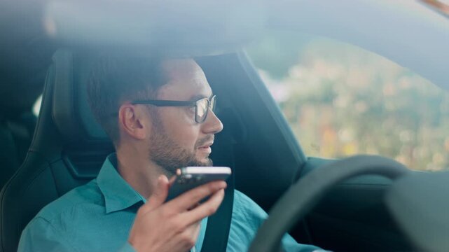 Serious bearded man in glasses recording voice message or using assistant on smartphone while sitting in car. Speaking clearly and staying composed. Handling communication during short roadside stop.