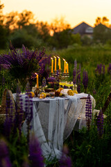 Romantic table decor for a loving couple on the blooming meadow with purple lupines. Two glasses of limoncello, flowers in a vase, candles, lemons, wooden furniture. Sunset, golden hour.