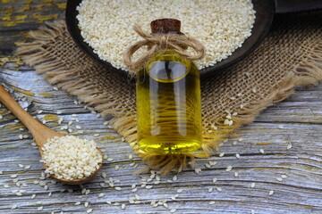 Glass bottle of sesame oil and raw sesame seeds in wooden spoon or bowl on rustic table. Uncooked...