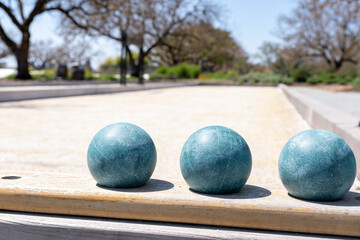 Three teal bocce balls lined up on a sunny court with trees and a walking path in the background at a springtime park