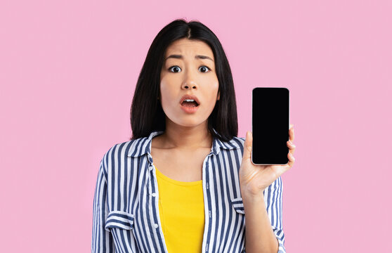 New App. Portrait of shocked amazed young asian woman holding smartphone with black blank screen in hand, showing device. Gadget with empty free space for mockup template. Technology Concept - Powered by Adobe
