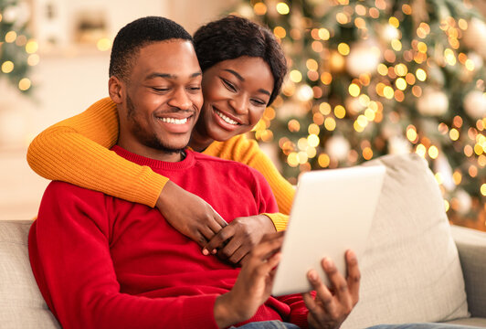 Congratulate family online during covid-19 pandemic. Happy millennial african american guy and lady hug and look at tablet and make video call in cozy living room interior with Xmas tree and garlands